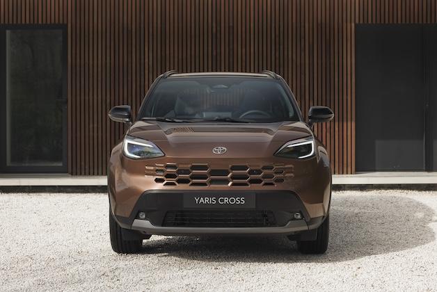 Nowa Toyota Yaris Cross w brązowym kolorze, widok z przodu, zaparkowana na jasnym żwirze przed budynkiem z drewnianą, lamelową elewacją.