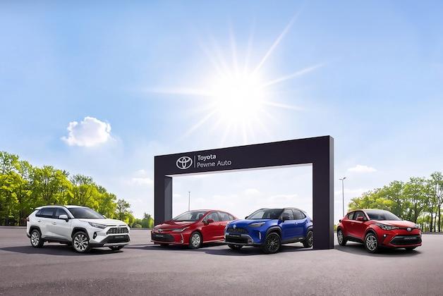 Cztery samochody marki Toyota (w tym modele RAV4, C-HR i Yaris Cross) zaparkowane na placu przed bramą z logo programu Toyota Pewne Auto w słoneczny dzień.