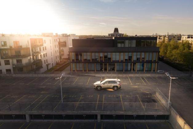 Jasna Toyota RAV4 zaparkowana na najwyższym poziomie wielopoziomowego parkingu na tle nowoczesnego budynku mieszkalnego o zachodzie słońca.