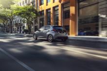 Szara Toyota Yaris jedzie miejską ulicą na tle nowoczesnych, pomarańczowo-brązowych budynków. Widok na tył i profil jadącego samochodu.