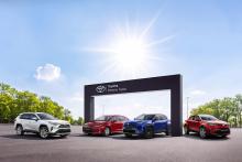 Cztery samochody marki Toyota (w tym modele RAV4, C-HR i Yaris Cross) zaparkowane na placu przed bramą z logo programu Toyota Pewne Auto w słoneczny dzień.